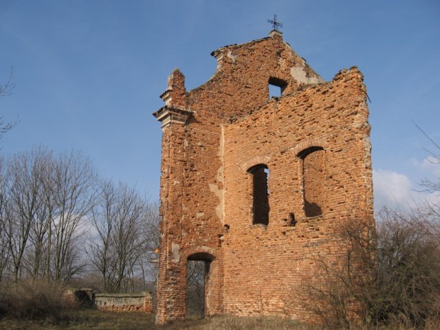 Ruiny ceglanego kościoła. Zachowana ściana frontowa z metalowym krzyżem na szczycie oraz fragment jednej z bocznych ścian z otworami po oknach.