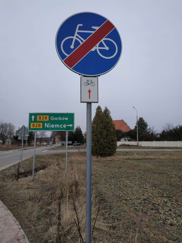 Znak drogowy koniec drogi dla rowerów, pod nim znak szlaku rowerowego: czarny rowerek a poniżej czerwona strzałka do góry