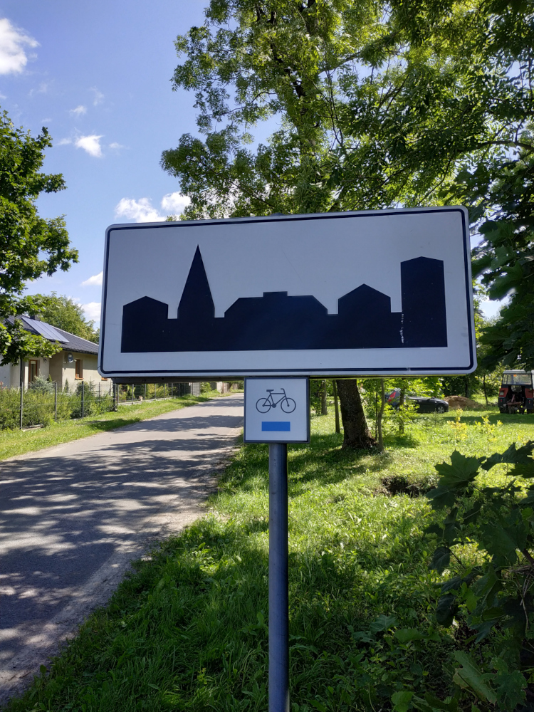 Znak drogowy teren zabudowany, pod nim znak szlaku rowerowego: czarny rowerek a poniżej niebieski pasek