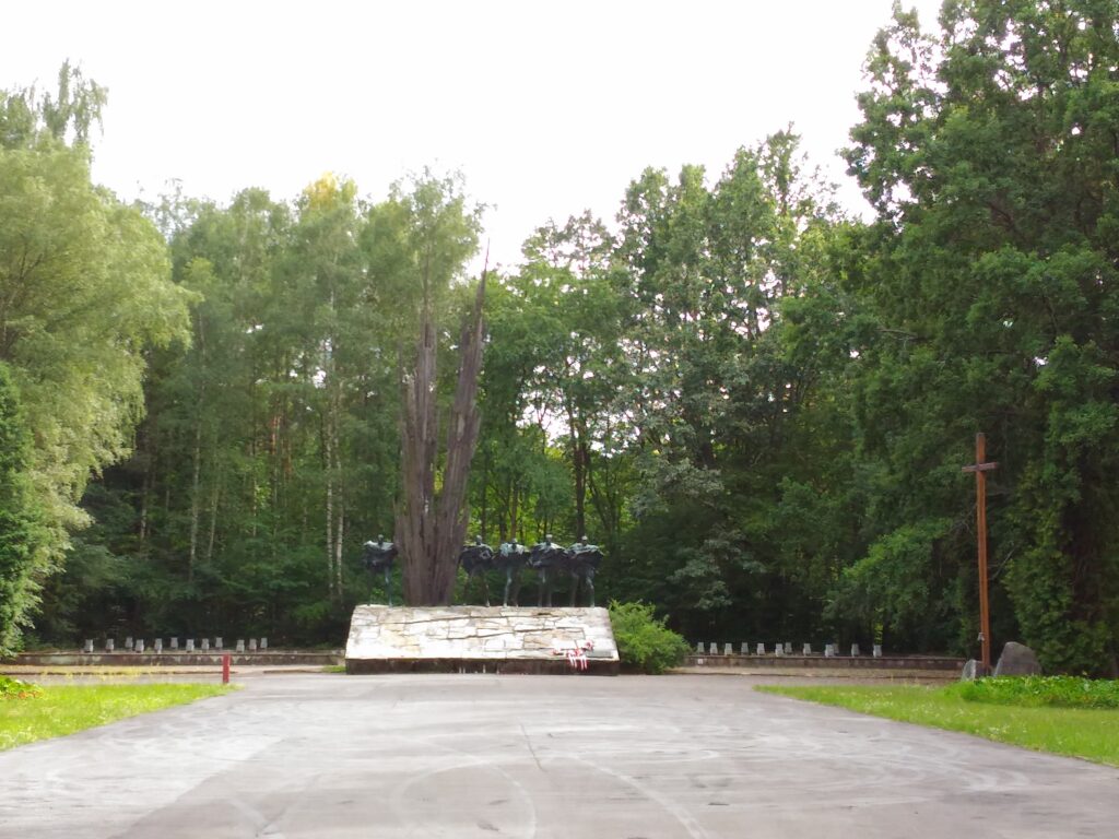 Betonowa szeroka droga a na jej końcu duży pomnik: pięć postaci żołnierzy, między nimi wysoko wtryskuje ziemia. Po prawej drewniany krzyż.