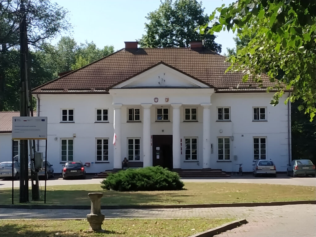 Biały pałac od frontu. Przed wejściem cztery gładkie wysokie kolumny. Kilka samochodów zaparkowanych przed wejściem