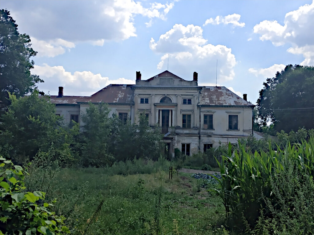 Zaniedbany spory pałac. Jasna elewacja, odpada tynk a z dachu farba. Na pierwszym planie zarośnięty trawnik