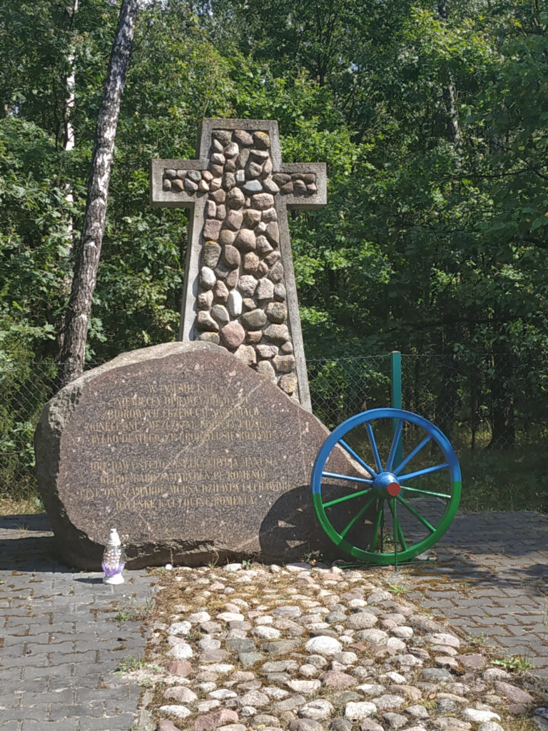 Pomnik: betonowy krzyż z kamieniami w środku, przed nim kamienna płyta z napisem z prawej niebiesko - zielone koło od dawnego wozu konnego.
