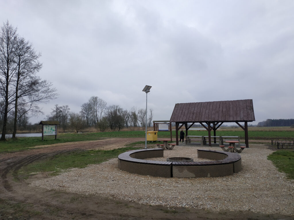 Miejsce ogniskowe otoczone ławkami a na drugim planie drewniana wiata turystyczna