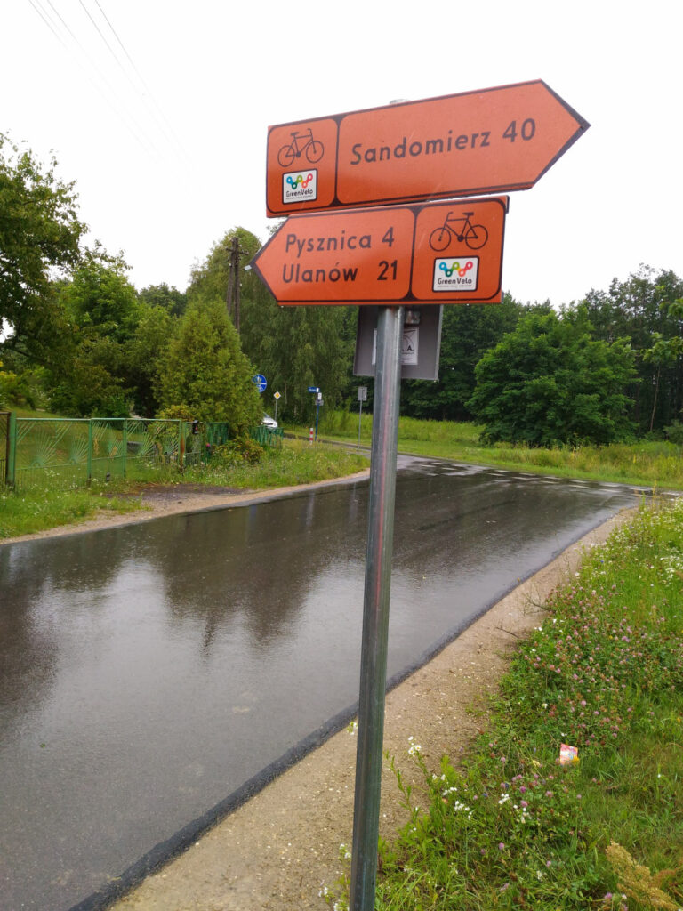 Dwa znaki szlaku rowerowego z pomarańczowym tłem. Górny kieruje w prawo i napis: Sandomierz 40, dolny kieruje w lewo i napis: Pysznica 4, Ulanów 21.