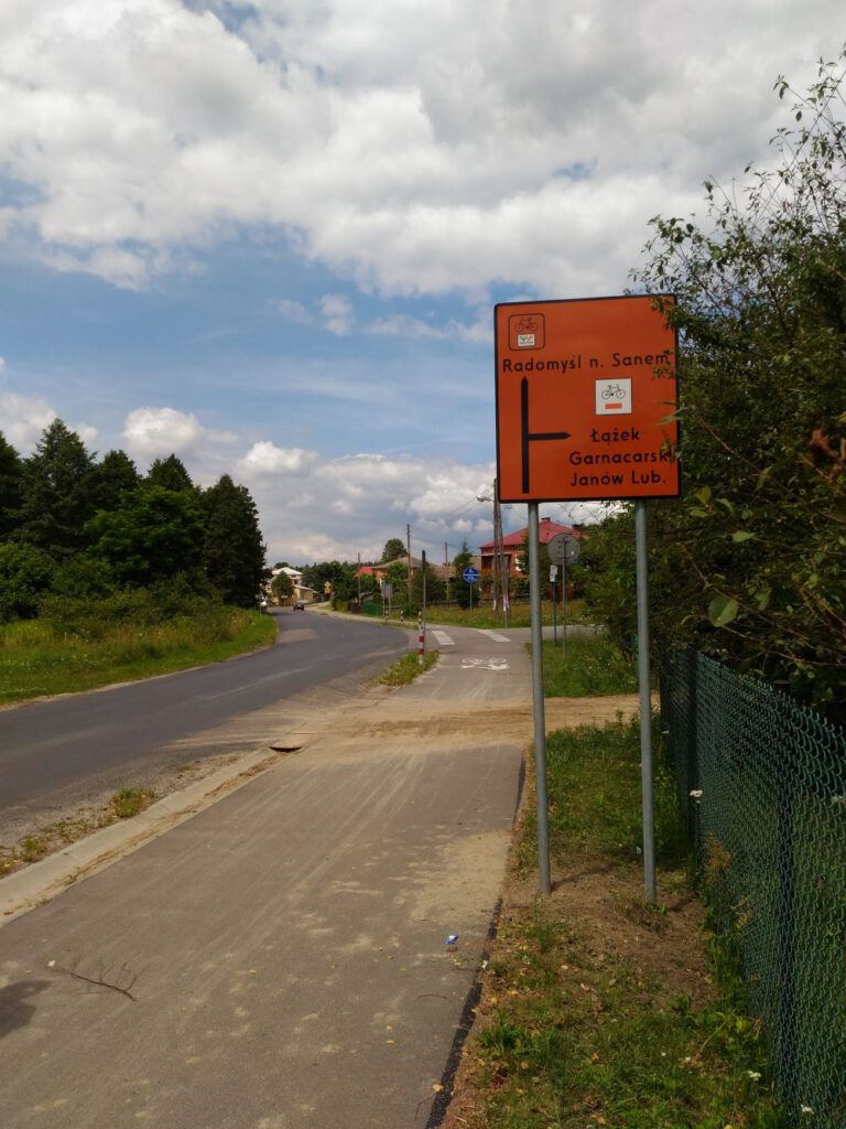 Znak szlaku rowerowego: pomarańczowy kwadrat. Z prawej strony znak szlaku rowerowego: czarny rower poniżej pomarańczowy prostokąt, a pod tym napis: Łążek Garncarski, Janów Lub. W plewym górnym rogu znak szlaku rowerowego: na górze czarny rowerek, poniżej logotyp szlaku Green Velo (pięć kolorowych kół), pod tym napis: Ulanów. W lewym dolnym rogu geometria skrzyżowania: czarna strzałka do góry oraz w prawo.