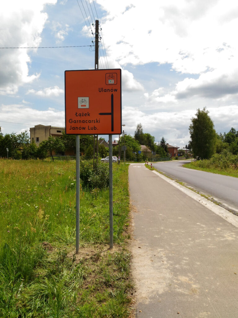 Znak szlaku rowerowego: pomarańczowy kwadrat. Z lewej strony znak szlaku rowerowego: czarny rower poniżej pomarańczowy prostokąt, a pod tym napis: Łążek Garncarski, Janów Lub. W prawym górnym rogu znak szlaku rowerowego: na górze czarny rowerek, poniżej logotyp szlaku Green Velo (pięć kolorowych kół), pod tym napis: Ulanów. W prawy dolnym rogu geometria skrzyżowania: czarna strzałka do góry oraz w prawo.