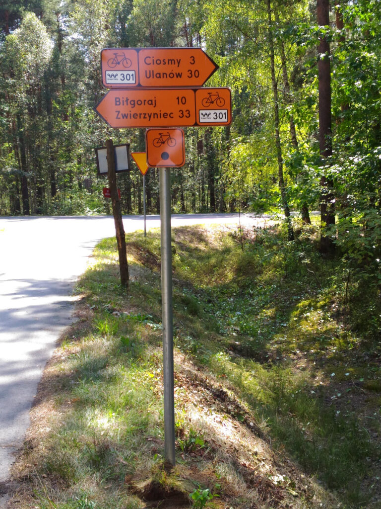 Trzy znaki szlaku rowerowego. Na górze prostokąt z pomarańczowym tłem, grot strzałki skierowany w prawo, z lewej czarny rowerek i biały prostokąt z liczbą 301, w prawej napis: Ciosmy 3, Ulanów 30. W środku prostokąt z pomarańczowym tłem, grot strzałki skierowany w lewo, z prawej czarny rowerek i biały prostokąt z liczbą 301, z lewej napis: Biłgoraj 10, Zwierzyniec 33. Poniżej kwadratowy znak z czarnym rowerkiem na górze i czarnym kołem niżej.