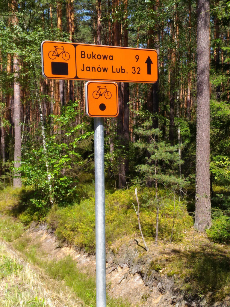 Dwa znaki szlaku rowerowego. Wyżej długi prostokąt z pomarańczowym tłem, z lewej czarny rowerek i czarny prostokącik, w prawej napis: Bukowa 9, Janów Lub. 32 i strzałka na wprost. Poniżej kwadratowy znak z czarnym rowerkiem na górze i czarnym kołem niżej.