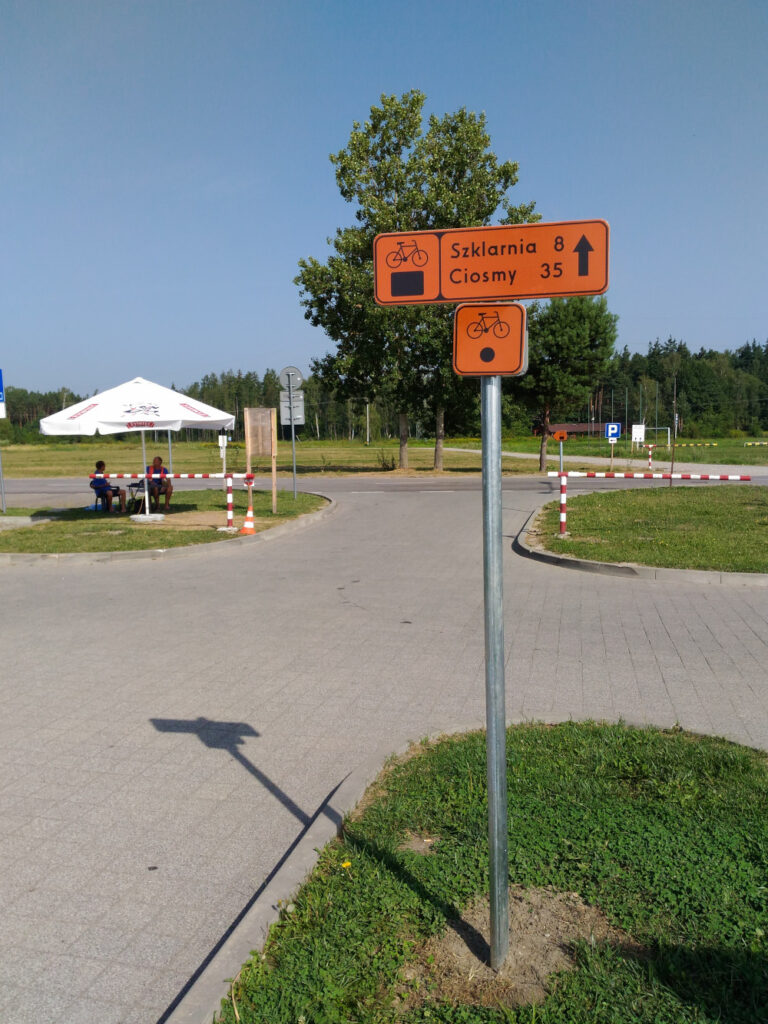 Dwa znaki szlaku rowerowego. Wyżej długi prostokąt z pomarańczowym tłem, z lewej czarny rowerek i czarny prostokącik, w prawej napis: Szklarnia 8, Ciosmy 35 i strzałka na wprost. Poniżej kwadratowy znak z czarnym rowerkiem na górze i czarnym kołem nizej.