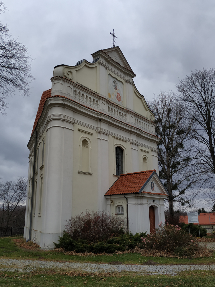Wejście i ściana boczna niedużego, jasnego kościoła. Na szczycie metalowy krzyż.