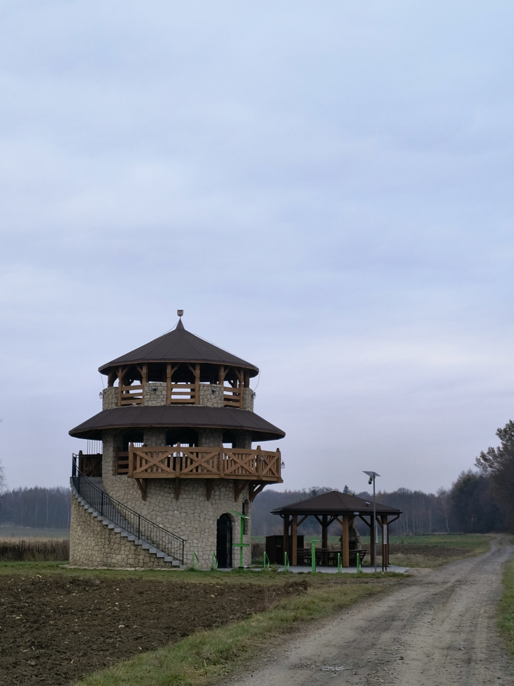 Kamienno - drewniana wieża widokowa. Z lewej na zewnątrz schody. Z prawej drewniana wiata. na pierwszym planie piaszczysta droga.