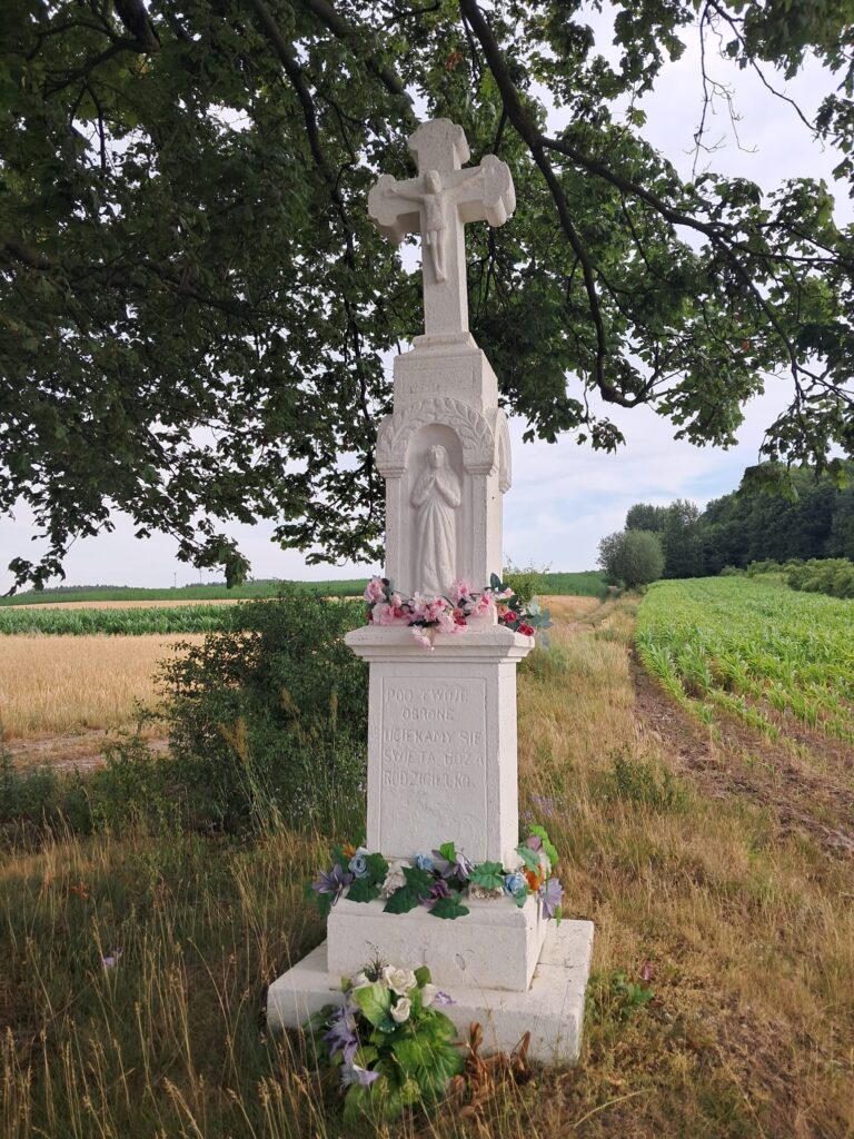Biała, kamienna kapliczka: na szczycie krzyż, poniżej figurka Maryi, na samym dole napis religijny. Wokół trawa, pola, gałęzie drzewa.