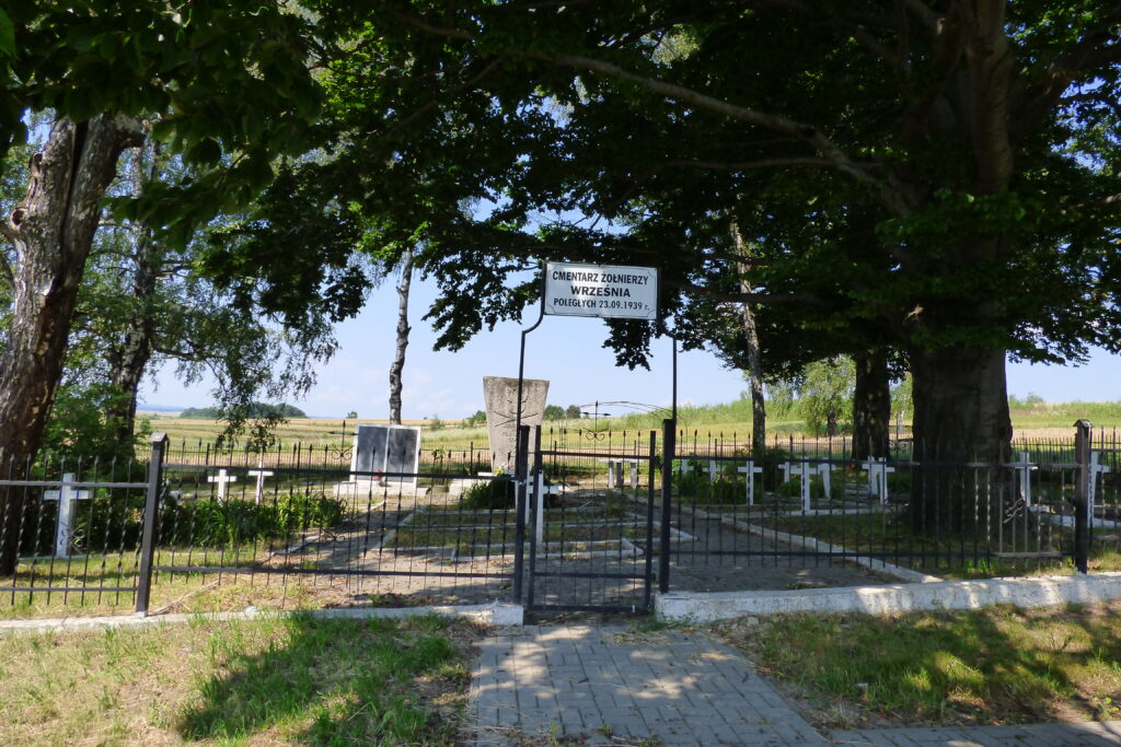 Otoczony metalowym płotem cmentarz. Nad wejściem biała tablica z napisem: cmentarz żołnierzy września poległych 23.09.1939r.