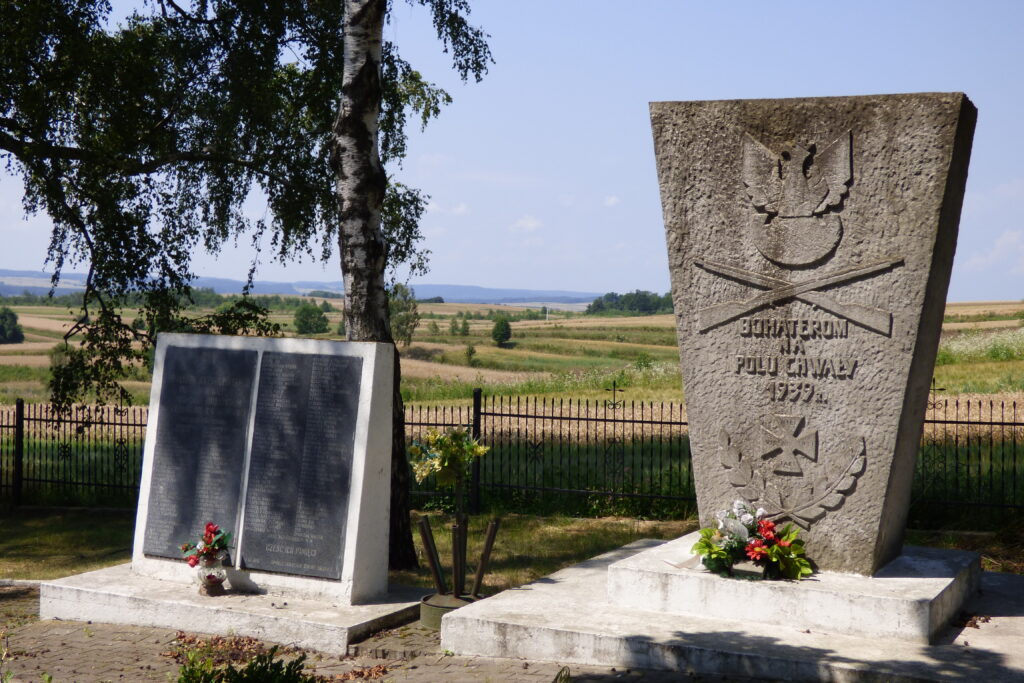 Dwa pomniki: z prawej kamienny z orłem, skrzyżowanymi dwoma karbinami i napisem: bohaterom na polu chwały 1939 r. Z lewej niższy pomnik, dwie czarne tablice z imionami i nazwiskami poległych żołnierzy.