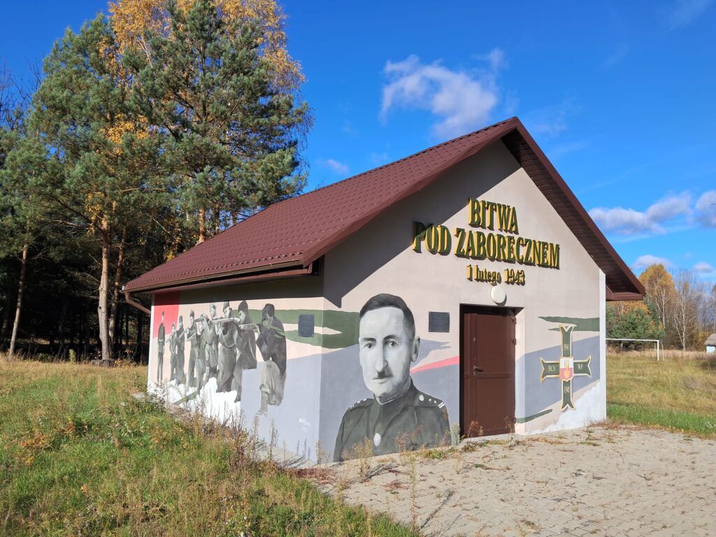 Nieduży budynek z brązowym dachem. Na ścianach żołnierze, partyzanci. na pierwszym planie portret ich dowódcy w mundurze polskiego oficera. Wokół łąka i drzewa.