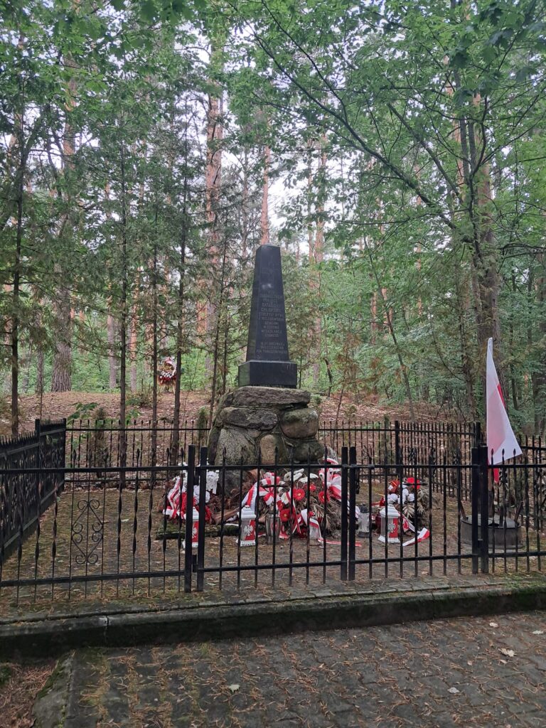 Metalowe ogrodzenie a za nim, na kamieniach, czarny pomnik z jasnymi, nieczytelnymi literami. Wokół las.